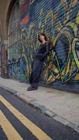 Retrato-De-Moda-Al-Aire-Libre-En-Video-Vertical-De-Una-Joven-Mujer-De-Estilo-Alternativo-Apoyada-Contra-Una-Pared-Cubierta-De-Graffitis-En-Una-Calle-De-La-Ciudad-De-Londres,-Reino-Unido,-En-Tiempo-Real